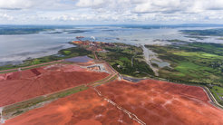 In the foreground, a section of the extensive area of a landfill site with rust-red mud, in the background a much smaller aluminium plant. The plant and landfill are located on a gulf, which can be seen in the upper half of the picture. Green meadows can be seen on the right.