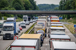 This picture, taken from a bridge over a motorway, shows heavy traffic with many lorries jammed on the two lanes in the direction of the camera. In the opposite direction, a few lorries and cars can be seen driving. A road bridge crosses the motorway a short distance away.
