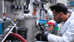 A man wearing a white coat and safety goggles is standing at the right-hand edge of the picture in front of an experimental plant, from which a vacuum drum and various operating instruments can be seen. He is looking through a window into the plant and grasping a control unit above his head.